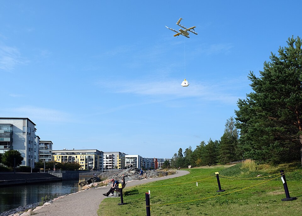 A Wing delivery drone in flight over a waterside park in Vuosaari, Helsinki, lowering a package on a tether, August 2021.